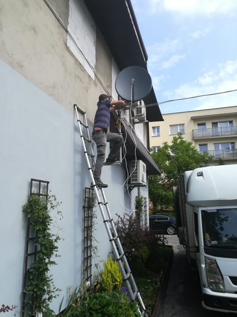 Instalator na drabinie montuje antenę satelitarną na ścianie budynku, widoczne kable i klimatyzator