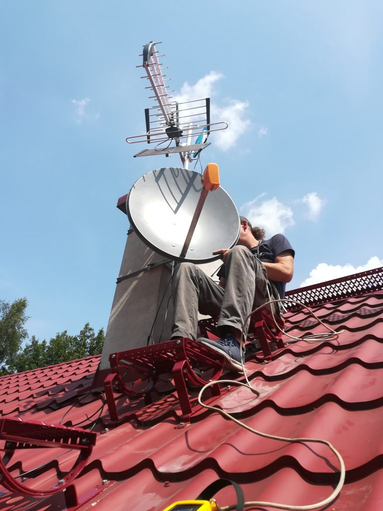 Praca na dachu: Instalator na czerwonej dachówce, na specjalnej platformie, montuje antenę satelitarną i antenę naziemną na kominie, widok z dołu na błękitne niebo.