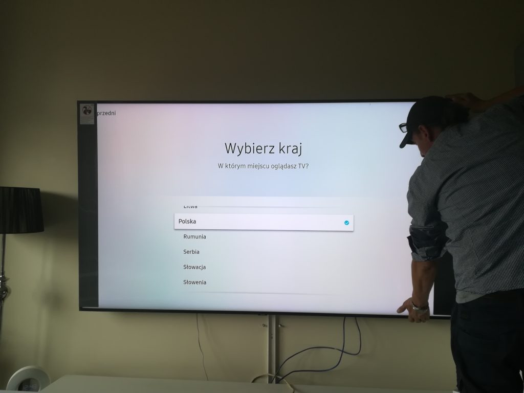 Mężczyzna w czapce instaluje duży telewizor, na ekranie menu wyboru kraju, widoczne kable i fragment lampy.