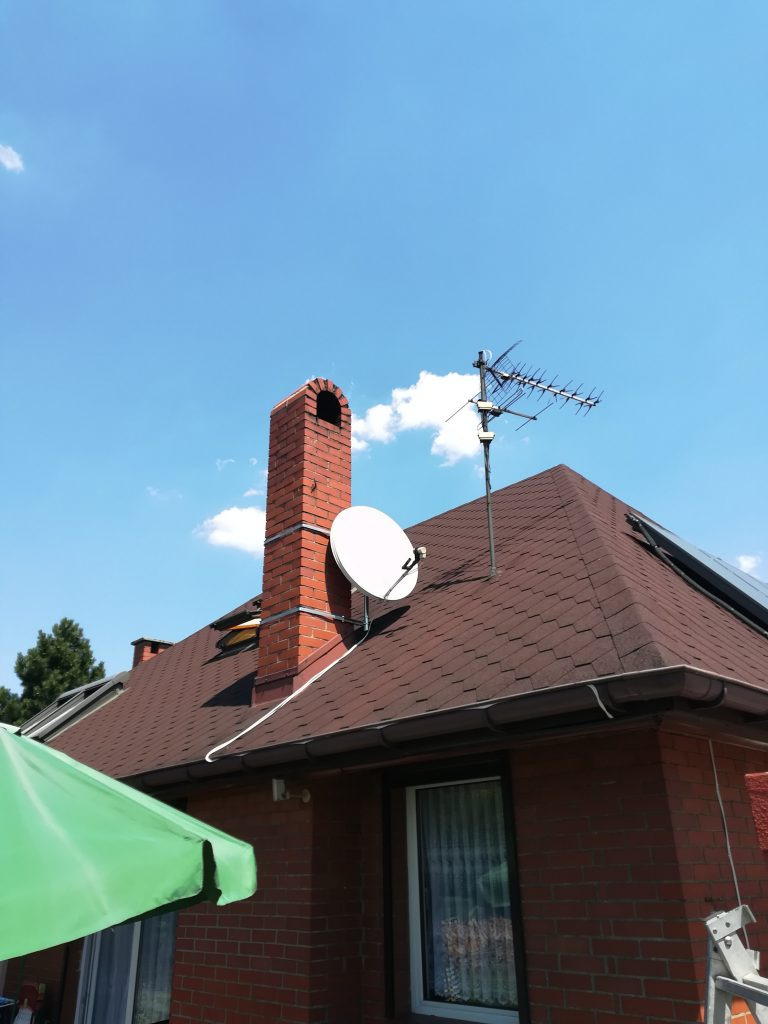 Antena telewizyjna i satelitarna zamontowane na dachu domu z ceglanym kominem, widok z dołu na tle błękitnego nieba.