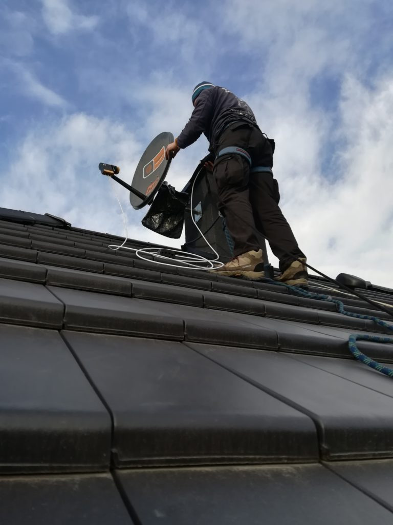 Pracownik w uprzęży na dachu z ciemnej dachówki, instaluje antenę satelitarną z pomarańczowym logo, widok z dołu na błękitne, zachmurzone niebo.