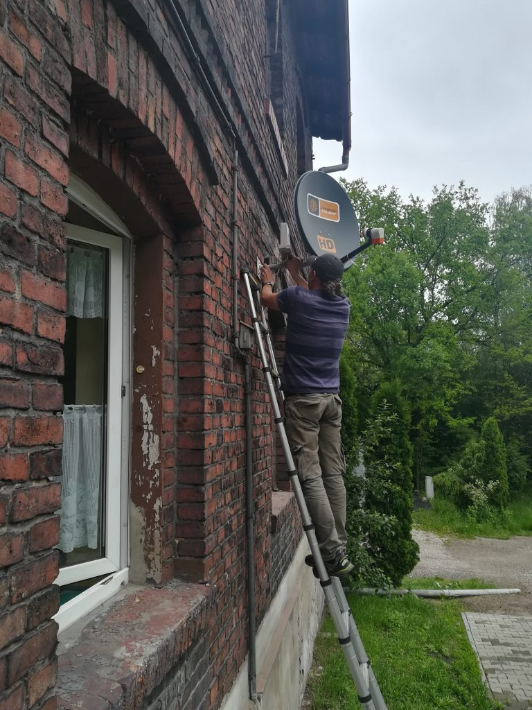Instalator na drabinie montuje antenę satelitarną HD na ceglanej ścianie budynku, widoczne okno z firanką i zieleń w tle.