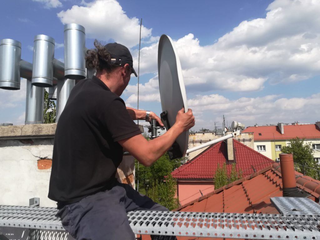 Instalator na dachu w Sosnowcu, trzymający antenę satelitarną na tle nieba i kominów wentylacyjnych.