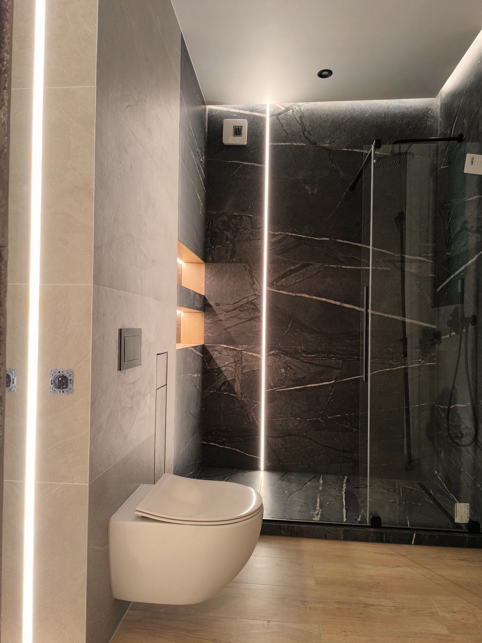 Nowoczesna łazienka z podwieszaną toaletą, kabiną prysznicową typu walk-in, marmurowymi płytkami w odcieniach szarości i oświetleniem LED wzdłuż ścian.