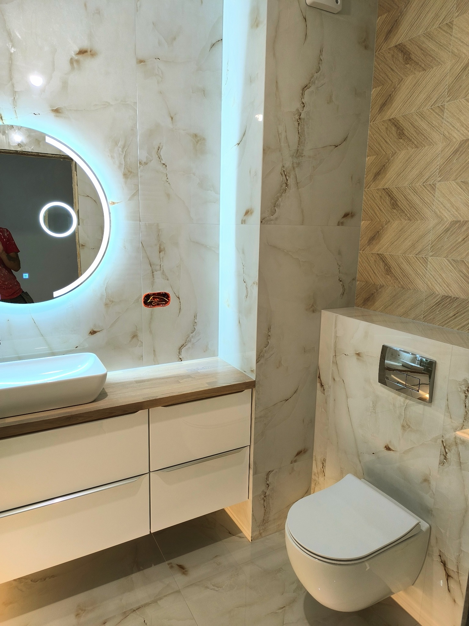 Nowoczesna łazienka z umywalką nablatową, okrągłym lustrem z podświetleniem LED, wiszącą toaletą i płytkami imitującymi marmur oraz drewno ułożonymi w jodełkę.