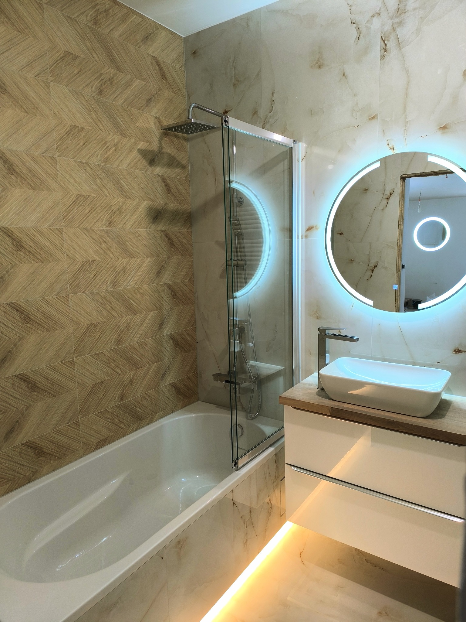 Nowoczesna łazienka z drewnopodobnymi płytkami ułożonymi w jodełkę na ścianie, marmurowe płytki na pozostałych ścianach, kabina prysznicowa ze szklaną ścianką, umywalka nablatowa z szafką...