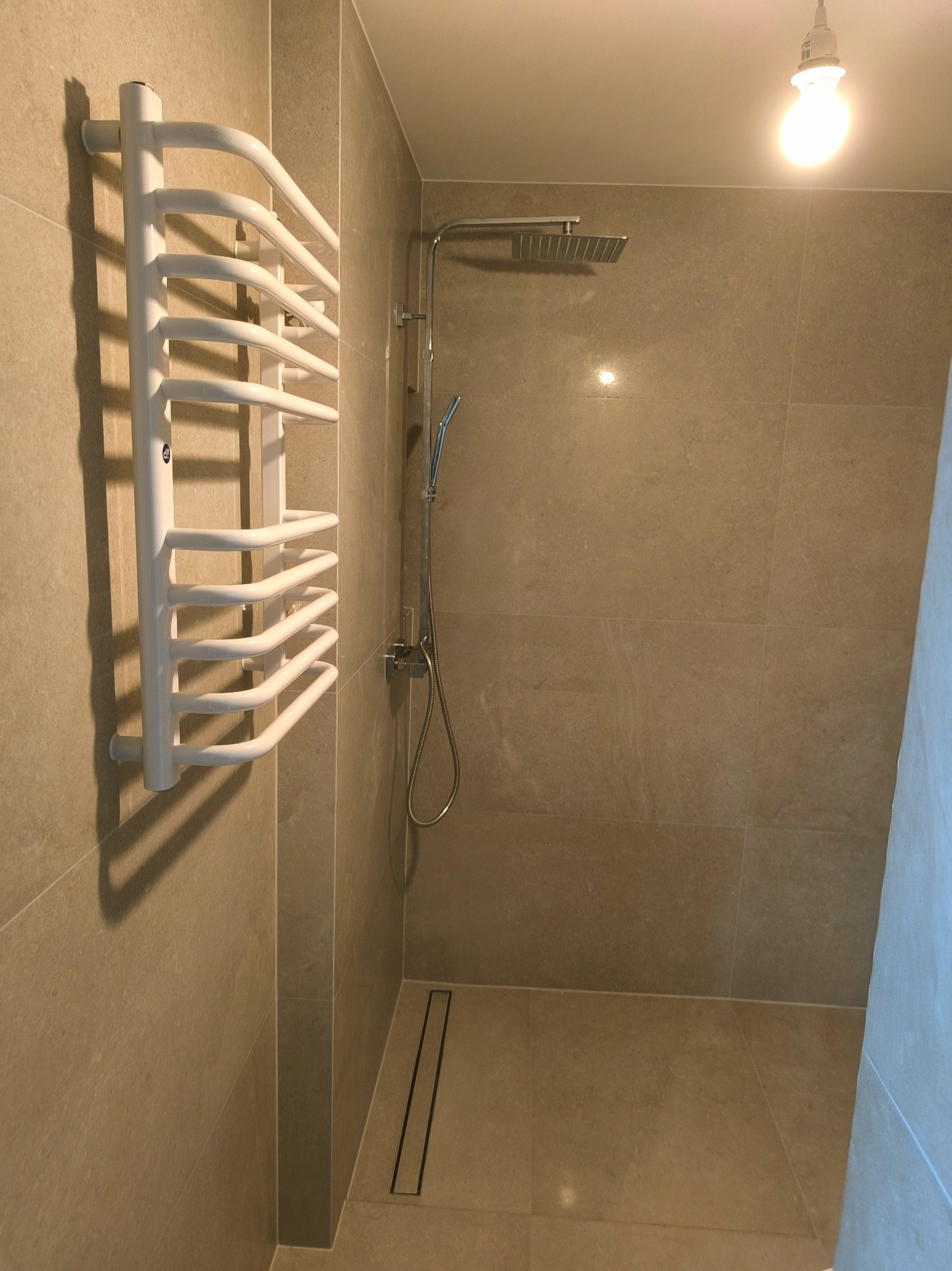 Nowoczesna kabina prysznicowa z odpływem liniowym i grzejnikiem drabinkowym w kolorze białym, wykończona płytkami w odcieniach beżu.