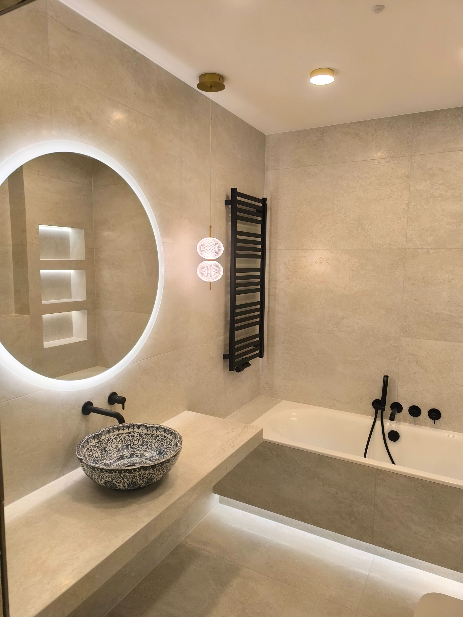 Jasna łazienka z beżowymi płytkami, okrągłe lustro z podświetleniem LED, dekoracyjna umywalka w stylu orientalnym, czarna armatura, grzejnik drabinkowy i oświetlenie wiszące.
