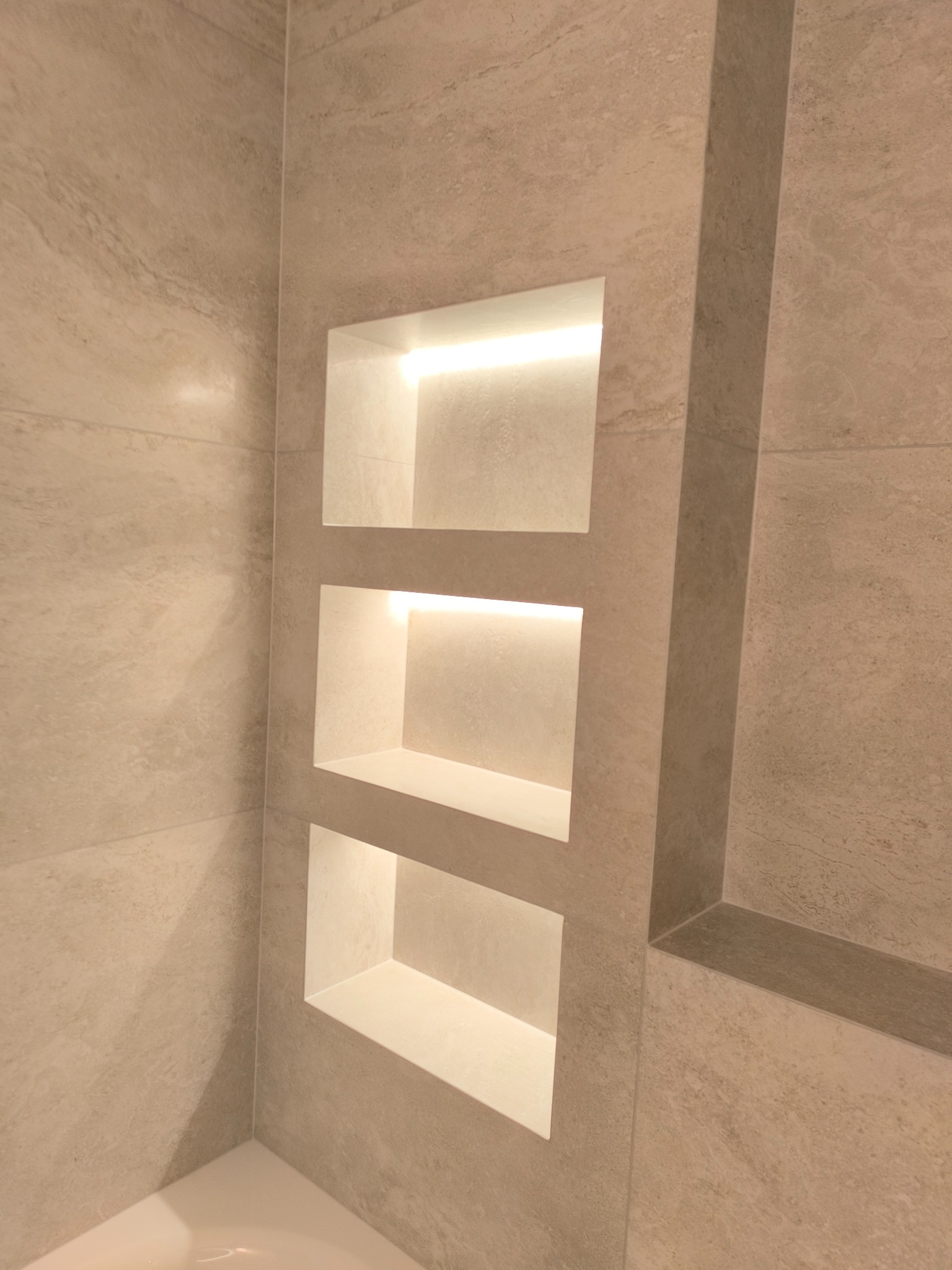 Beżowa łazienka z trzema podświetlanymi wnękami w ścianie wyłożonej płytkami imitującymi kamień.