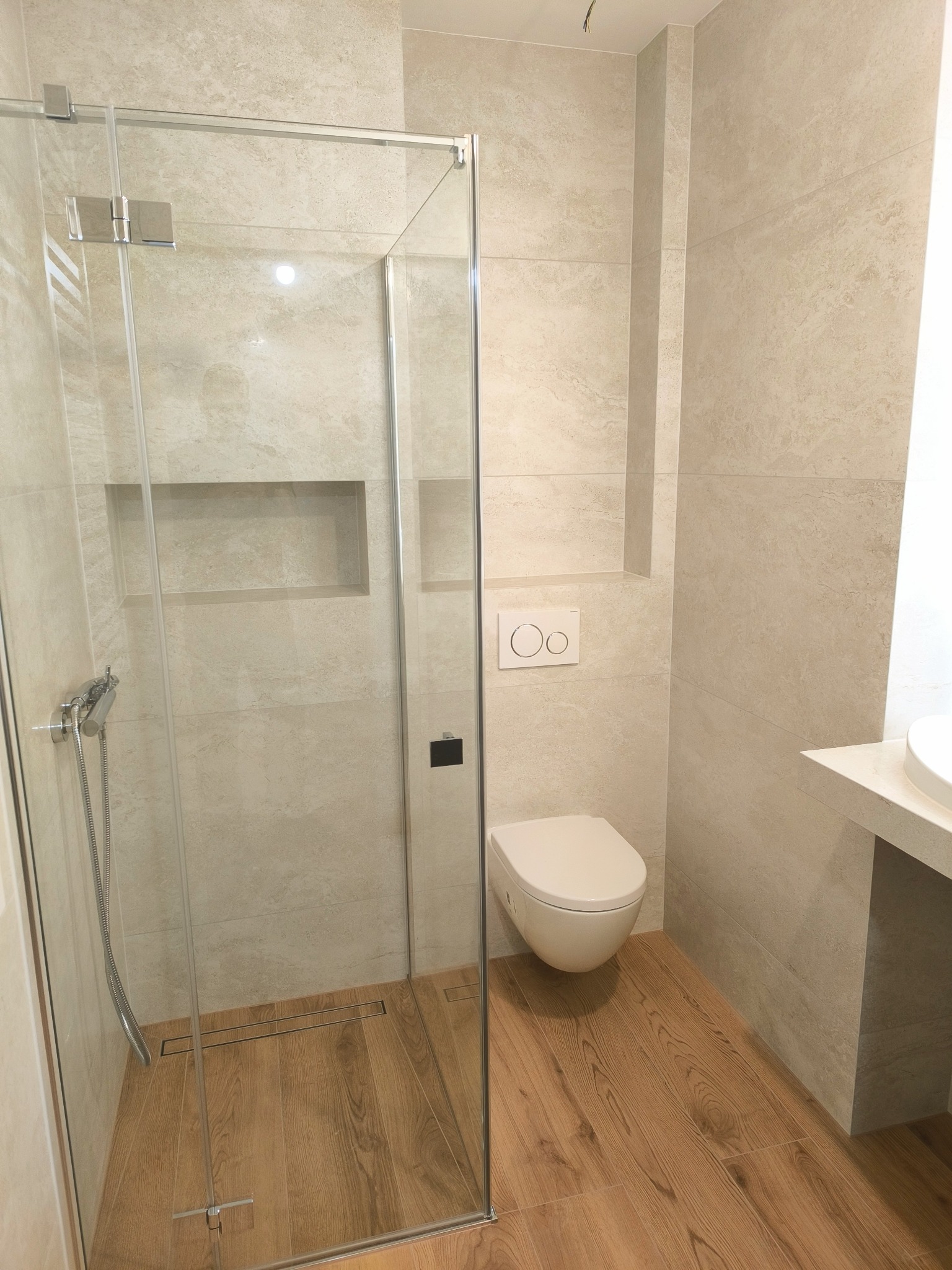 Nowoczesna łazienka z kabiną prysznicową typu walk-in, wykończona beżowymi płytkami imitującymi kamień i drewnopodobną podłogą, z widocznym odpływem liniowym.