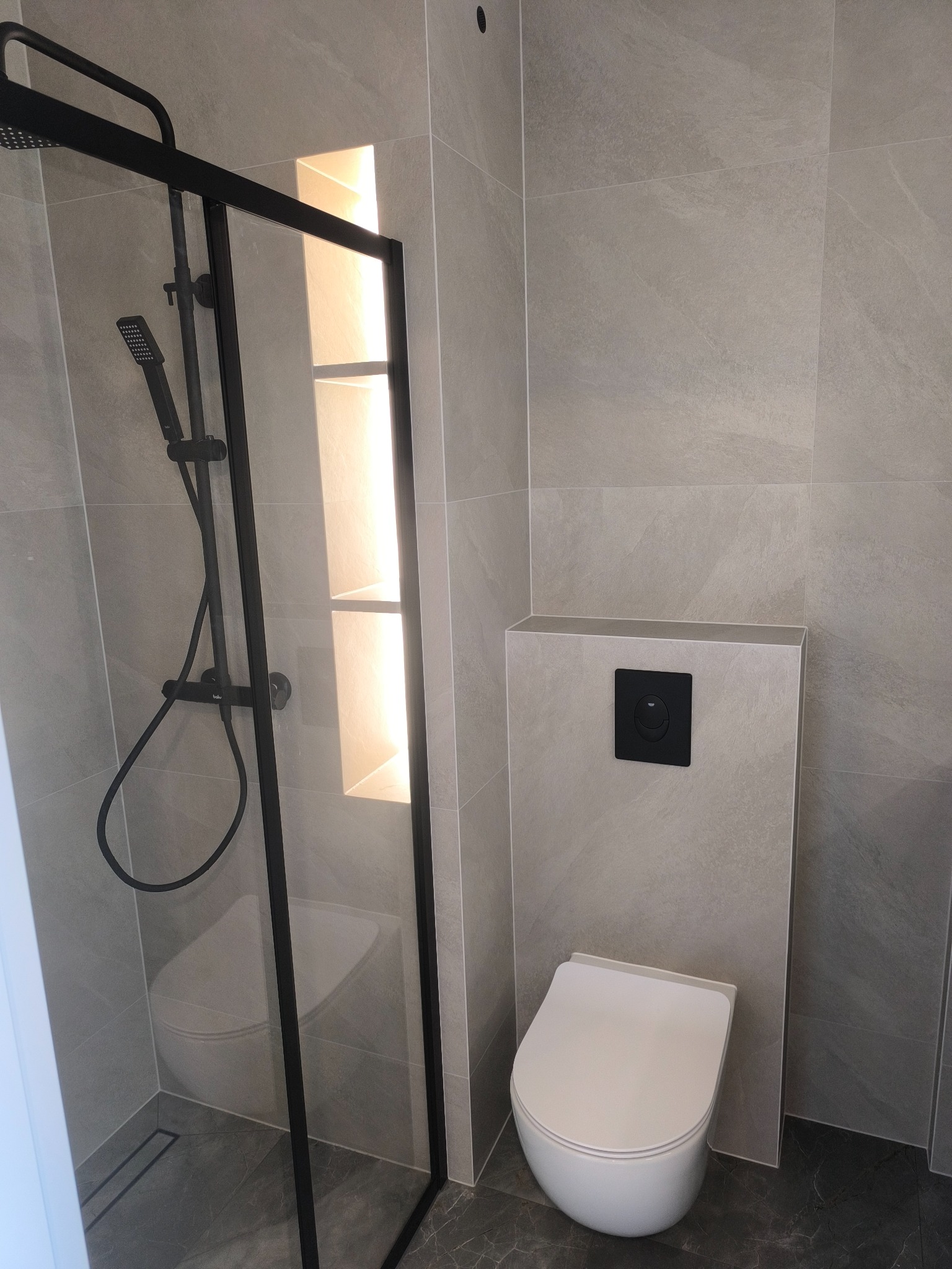 Nowoczesna łazienka z szarymi płytkami imitującymi kamień, kabina prysznicowa z czarną ramą, podtynkowa toaleta z czarnym przyciskiem spłukiwania i wnęka z podświetleniem.