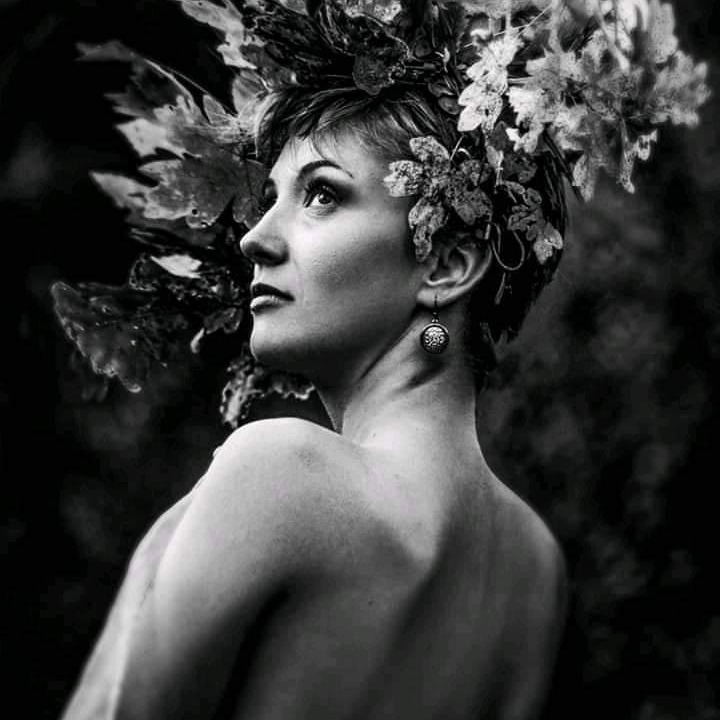 Czarno-białe portretowe ujęcie kobiety z liśćmi we włosach, spoglądającej w górę, z widocznym kolczykiem i nagimi ramionami na ciemnym tle.