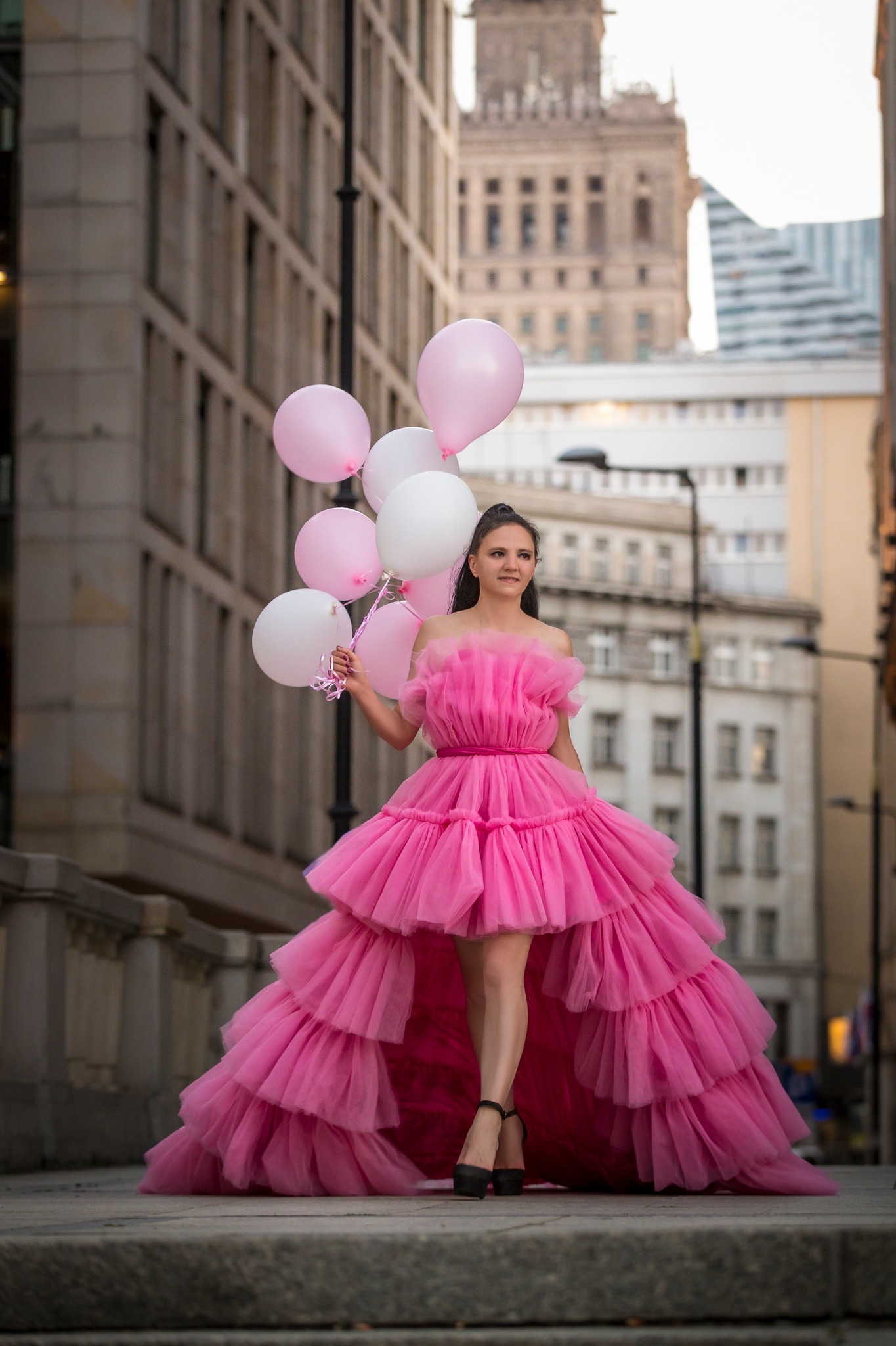 Kobieta w tiulowej, różowej sukni pozuje z białymi i różowymi balonami na tle miejskiej architektury, w tym Pałacu Kultury.
