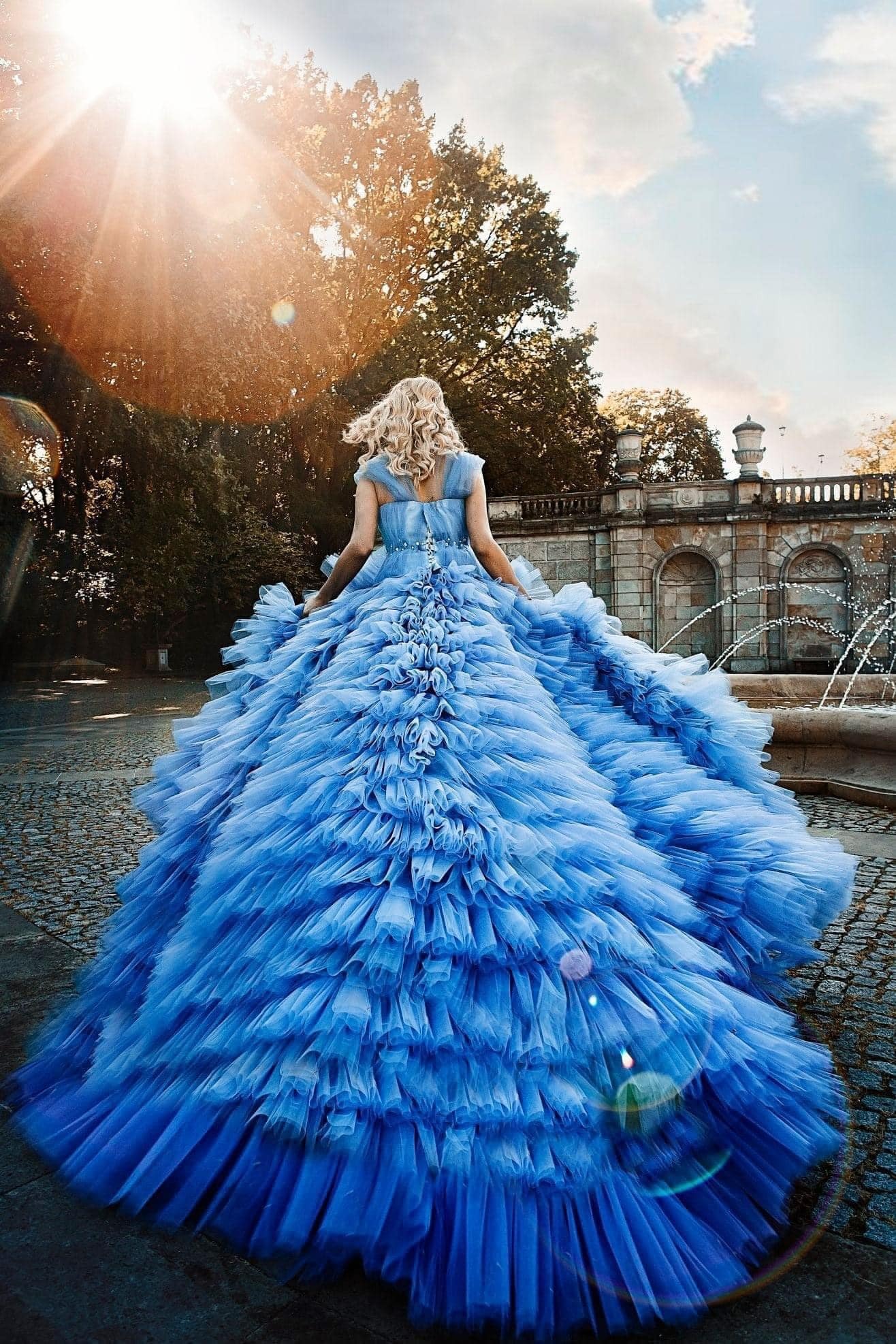 Kobieta w błękitnej, balowej sukni z falbanami, sfotografowana od tyłu w parku w Warszawie, idąca w kierunku fontanny i kamiennego muru w słoneczny dzień.