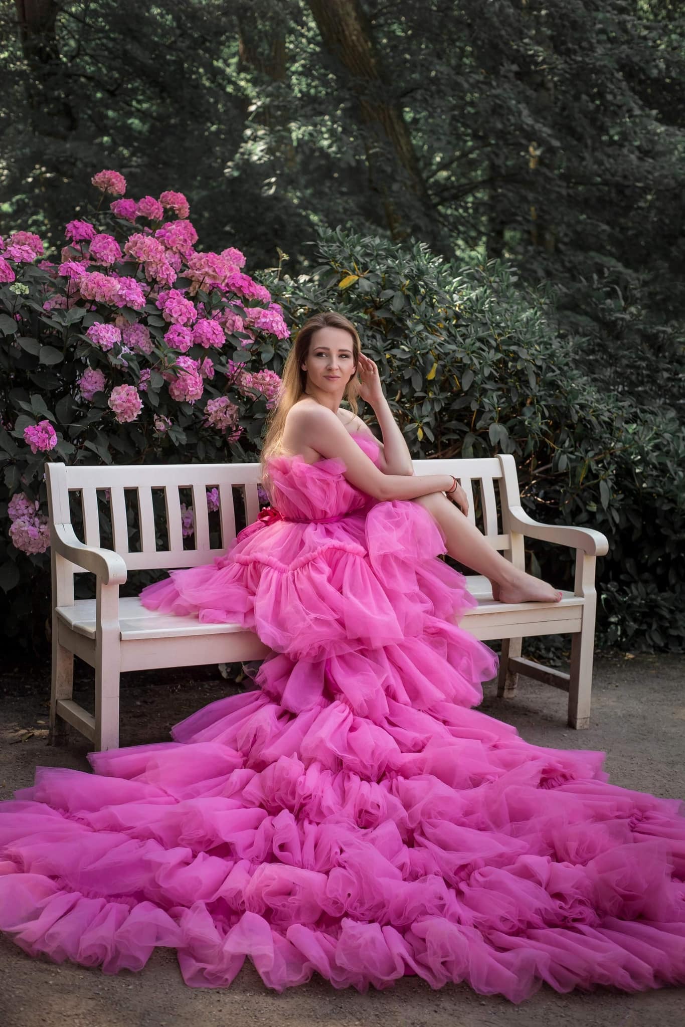Kobieta w obszernej, różowej sukni z tiulu siedzi na białej ławce w ogrodzie, obok kwitnących hortensji.