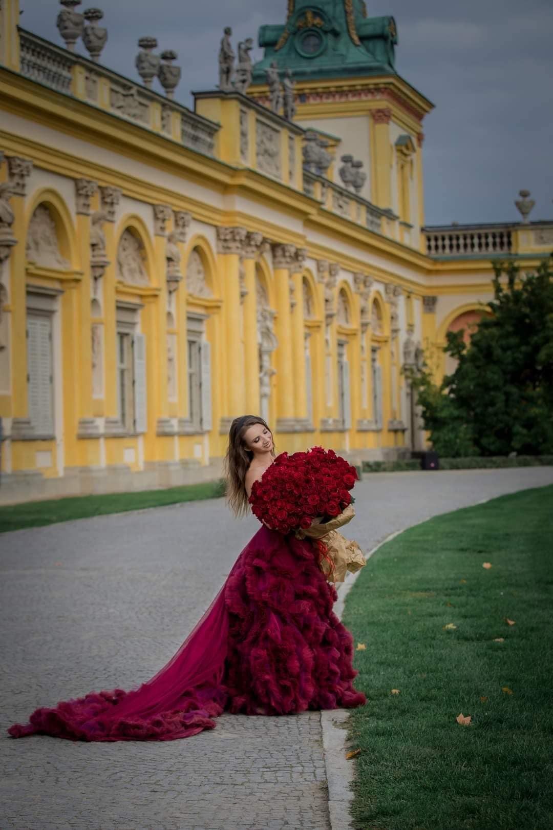 Kobieta w bordowej sukni z długim trenem, trzymająca ogromny bukiet czerwonych róż na tle pałacowej architektury z kolumnami.