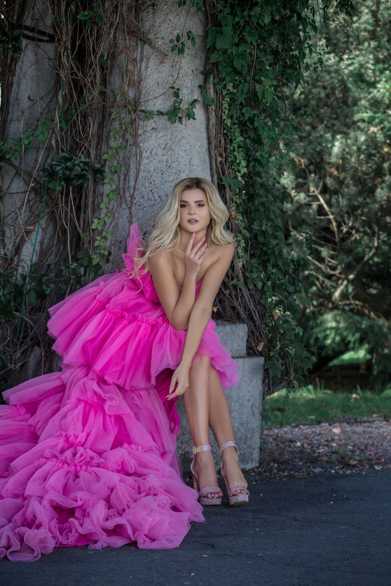 Kobieta w tiulowej sukni w odcieniu fuksji, pozująca na tle kamiennej konstrukcji porośniętej bluszczem, z delikatnym makijażem i blond włosami.