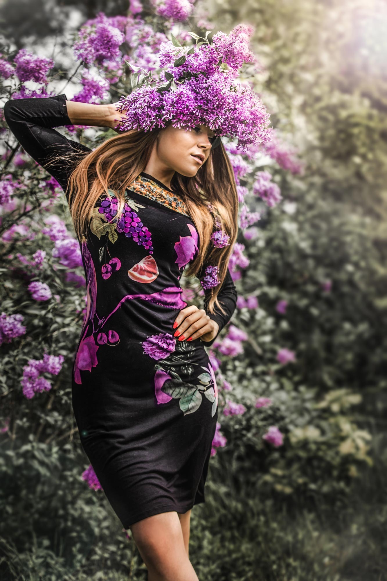 Kobieta w czarnej sukience z kwiatowym wzorem, z wiankiem z fioletowych kwiatów na głowie, pozuje na tle kwitnących krzewów.
