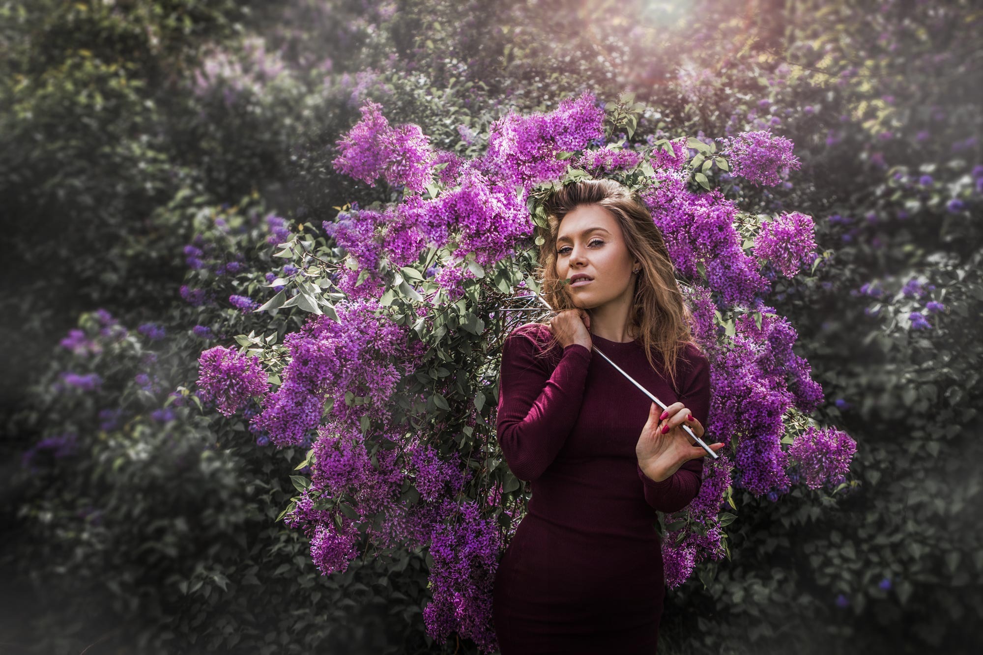 Portret kobiety w bordowej sukience, trzymającej metalowy wieszak na ubrania na tle kwitnącego krzewu bzu z fioletowymi kwiatami.