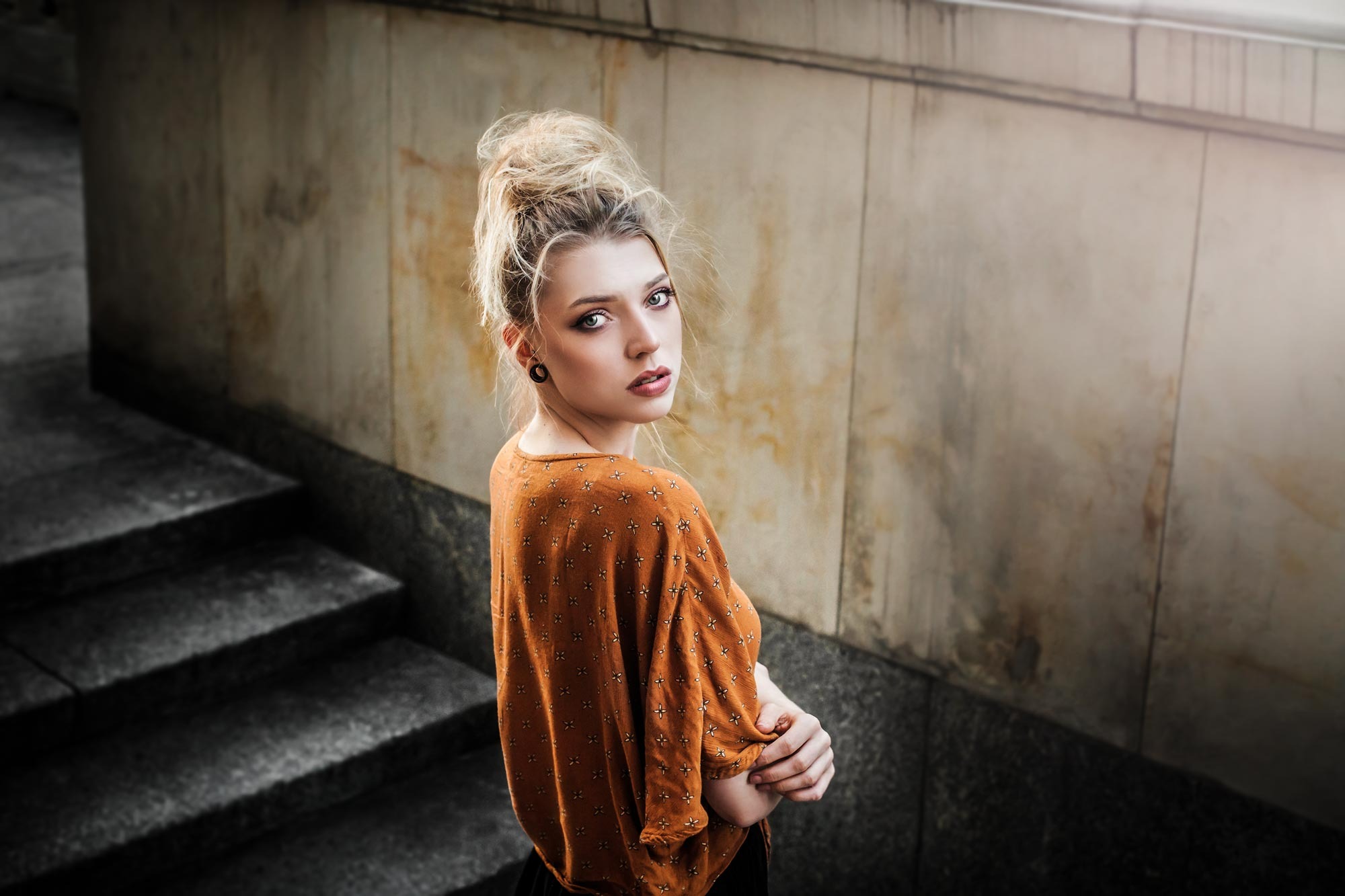Portret kobiety z wysoko upiętymi blond włosami, w pomarańczowej bluzce w drobne wzory, spoglądającej przez ramię na tle marmurowej ściany i schodów.