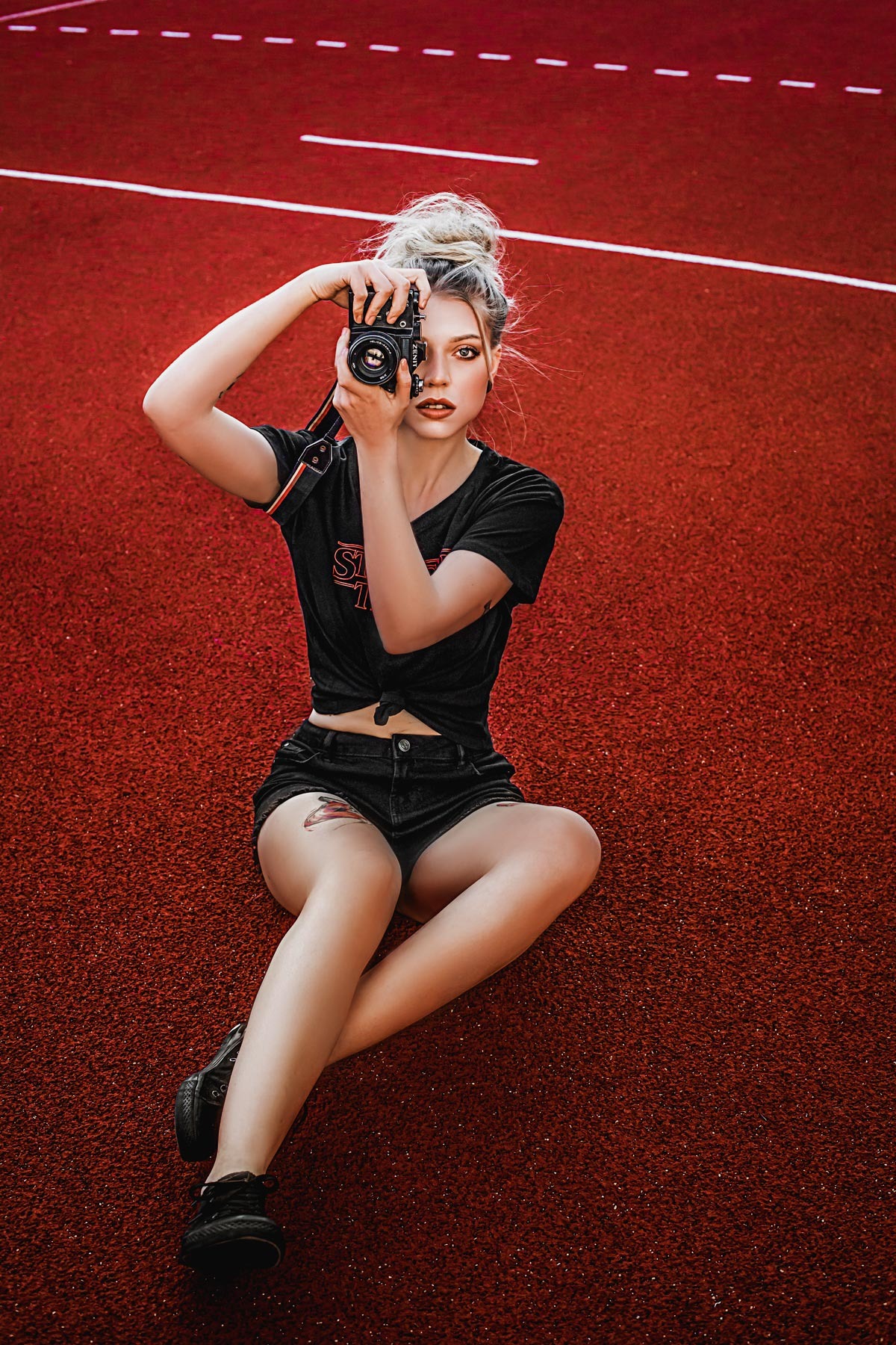 Młoda kobieta z blond włosami w koku siedzi na czerwonym tartanowym boisku, trzymając w dłoniach aparat fotograficzny Zenit i celując obiektywem w widza. Ubrana w czarny t-shirt i szorty,...