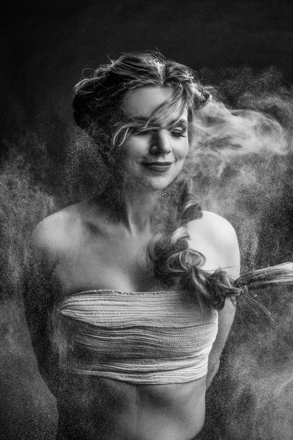 Portret kobiety w czerni i bieli, otoczonej chmurą białego pyłu, z warkoczem opadającym na ramię i bandażem na torsie, w klimacie artystycznym.