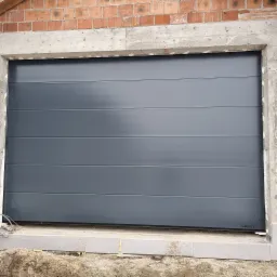 Szara segmentowa konstrukcja w trakcie montażu w surowym otworze garażowym z widocznymi elementami muru i betonu.