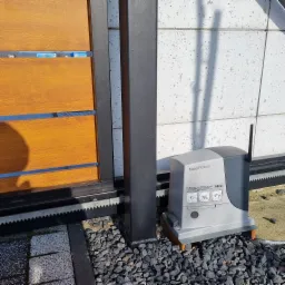 Zewnętrzny napęd Nice Robus zamontowany przy przesuwnej bramie wjazdowej z drewnianymi panelami, widoczne elementy montażowe i zębatka, tło stanowi betonowa ściana.