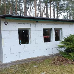 Ocieplony budynek gospodarczy z trzema oknami, styropianowe bloki z widocznymi otworami montażowymi, w tle las iglasty, widoczna puszka pianki montażowej przy oknie.