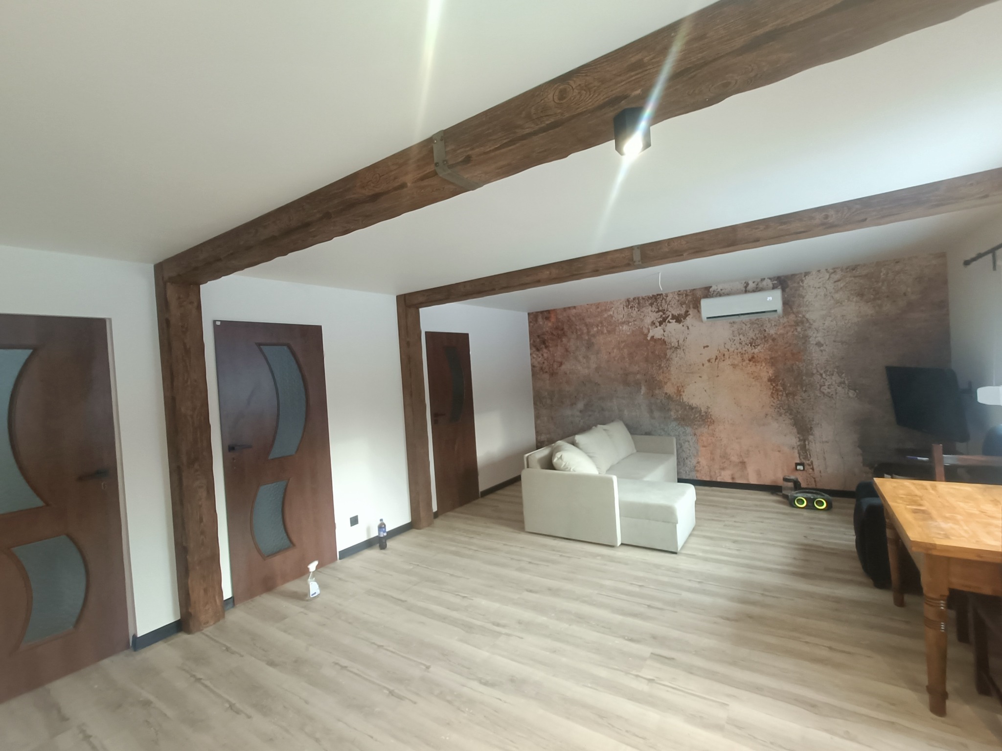 Jasne wnętrze po remoncie: drewniane belki stropowe, podłoga panelowa, ściana z efektem rdzy, biała sofa. Widoczne drzwi i klimatyzacja. Nowoczesny design.