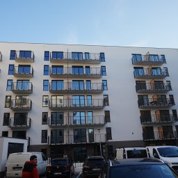 G&M+ sp. z o.o. - Nowoczesny budynek wielorodzinny z balkonami, jasna elewacja, widok z dołu, zaparkowane samochody i dostawcze busy, błękitne niebo w tle.