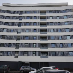 G&M+ sp. z o.o. - Nowoczesny budynek wielorodzinny z białymi, falującymi balkonami i szarymi pasami między oknami. Na dole widoczne zaparkowane samochody.