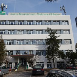 G&M+ sp. z o.o. - Biały budynek biurowy z szarymi akcentami okiennymi i logo Agencji Restrukturyzacji i Modernizacji Rolnictwa na dachu, otoczony drzewami i samochodami na parkingu.