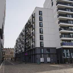 G&M+ sp. z o.o. - Nowoczesny budynek mieszkalny z balkonami, widoczny z perspektywy ulicy wyłożonej kostką. Kontrastujące kolory elewacji: biel i szarość. W tle starsza kamienica.