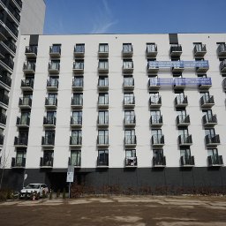 G&M+ sp. z o.o. - Nowoczesny budynek wielorodzinny w Łodzi z balkonami i szarą elewacją. Na niektórych balkonach wiszą banery reklamowe. Widok z dołu, w tle błękitne niebo.