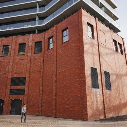 G&M+ sp. z o.o. - Ceglana fasada budynku z nowoczesną nadbudową. Widoczne okna, kratki wentylacyjne i chodnik z kostki. Osoba w oddali dodaje skali budowli. Architektura industrialna.