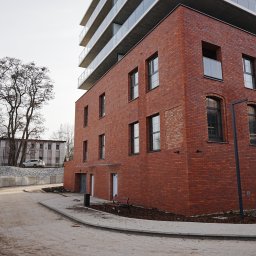 G&M+ sp. z o.o. - Ceglana elewacja budynku z oknami, widok z poziomu chodnika. W tle drzewa i fragment drugiego budynku. Nowoczesna architektura z elementami tradycyjnymi.