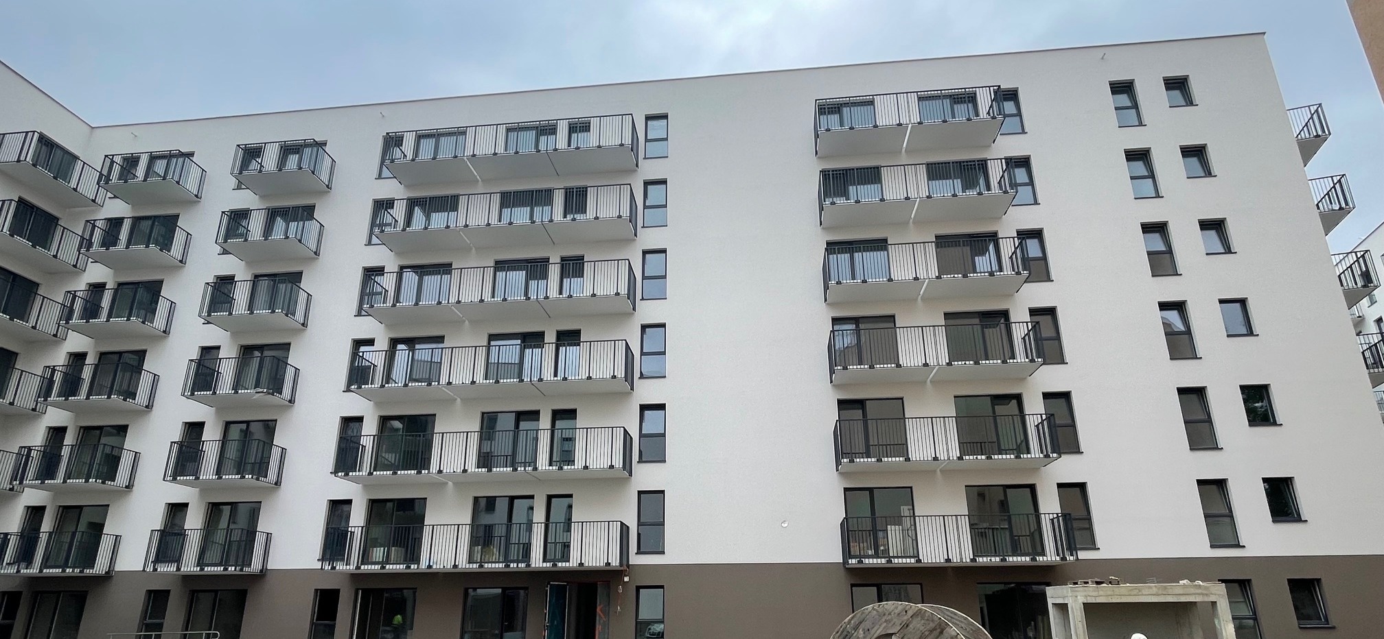 Nowoczesny budynek wielorodzinny z białymi elewacjami i ciemnymi balkonami, widok z dołu, fragment placu budowy w tle.