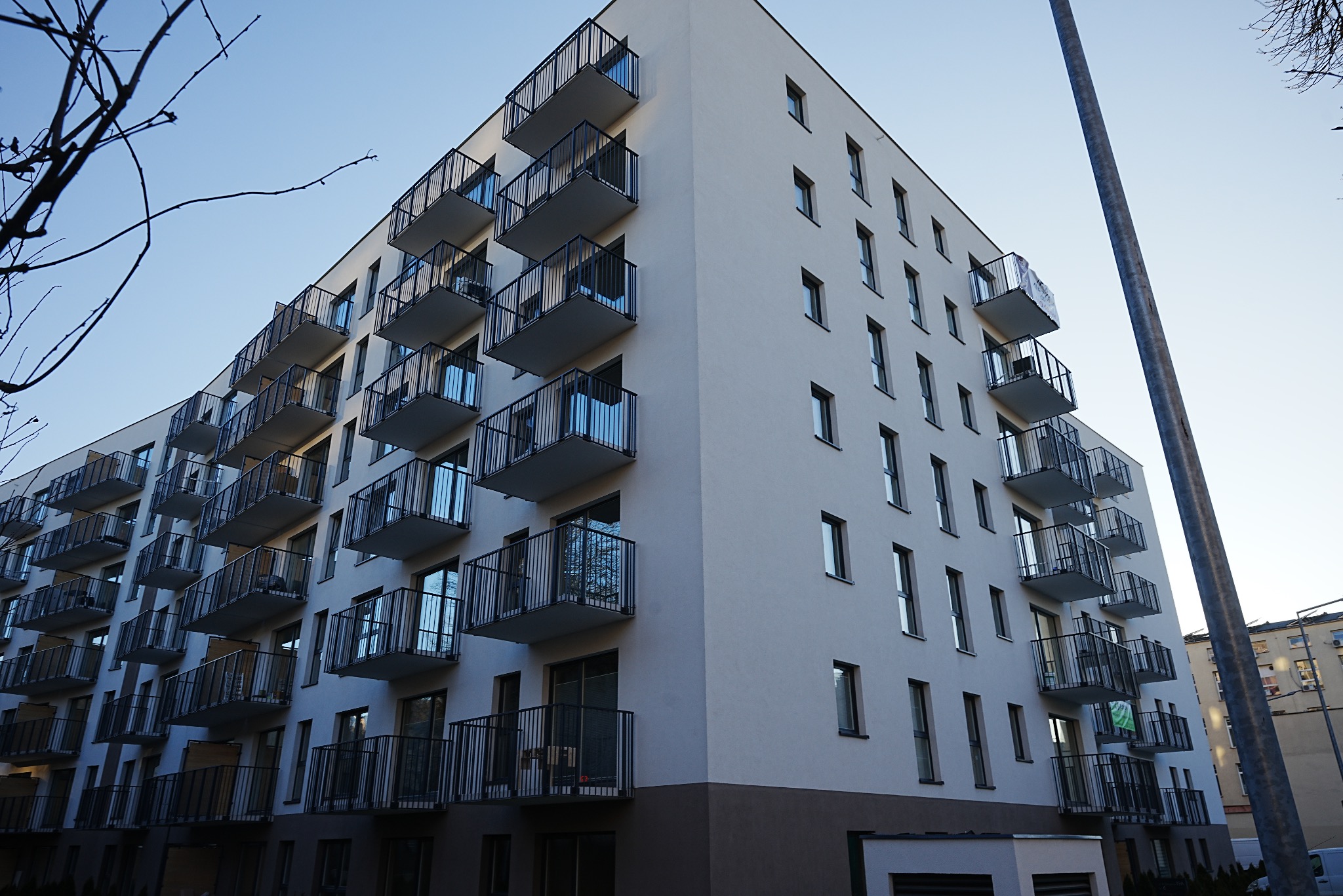 Nowoczesny budynek wielorodzinny z białymi ścianami i balkonami z czarnymi balustradami, widziany pod kątem z dołu, na tle błękitnego nieba.