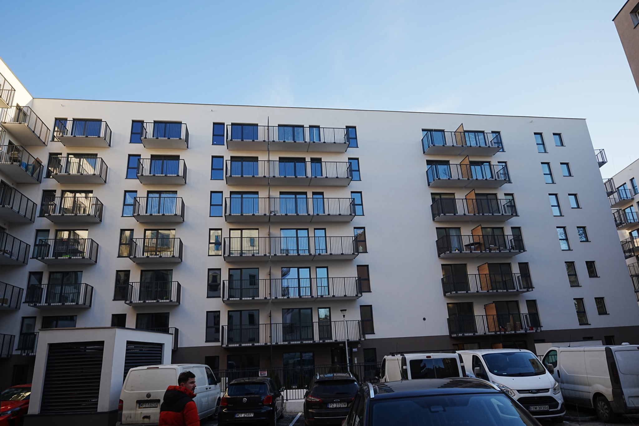 Nowoczesny budynek wielorodzinny z balkonami, jasna elewacja, widok z dołu, zaparkowane samochody i dostawcze busy, błękitne niebo w tle.
