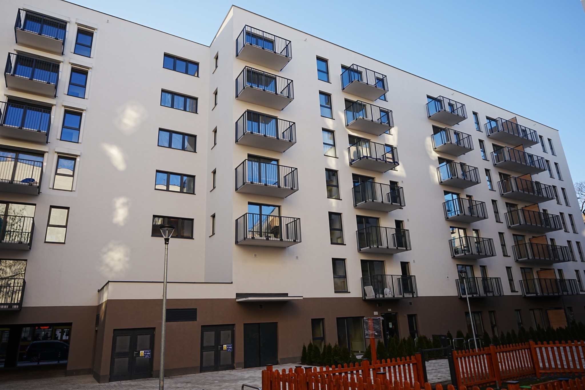 Nowoczesny budynek wielorodzinny z balkonami, wykończony w jasnych barwach z ciemnobrązowym pasem na dole. Widoczne detale architektoniczne i oświetlenie zewnętrzne.