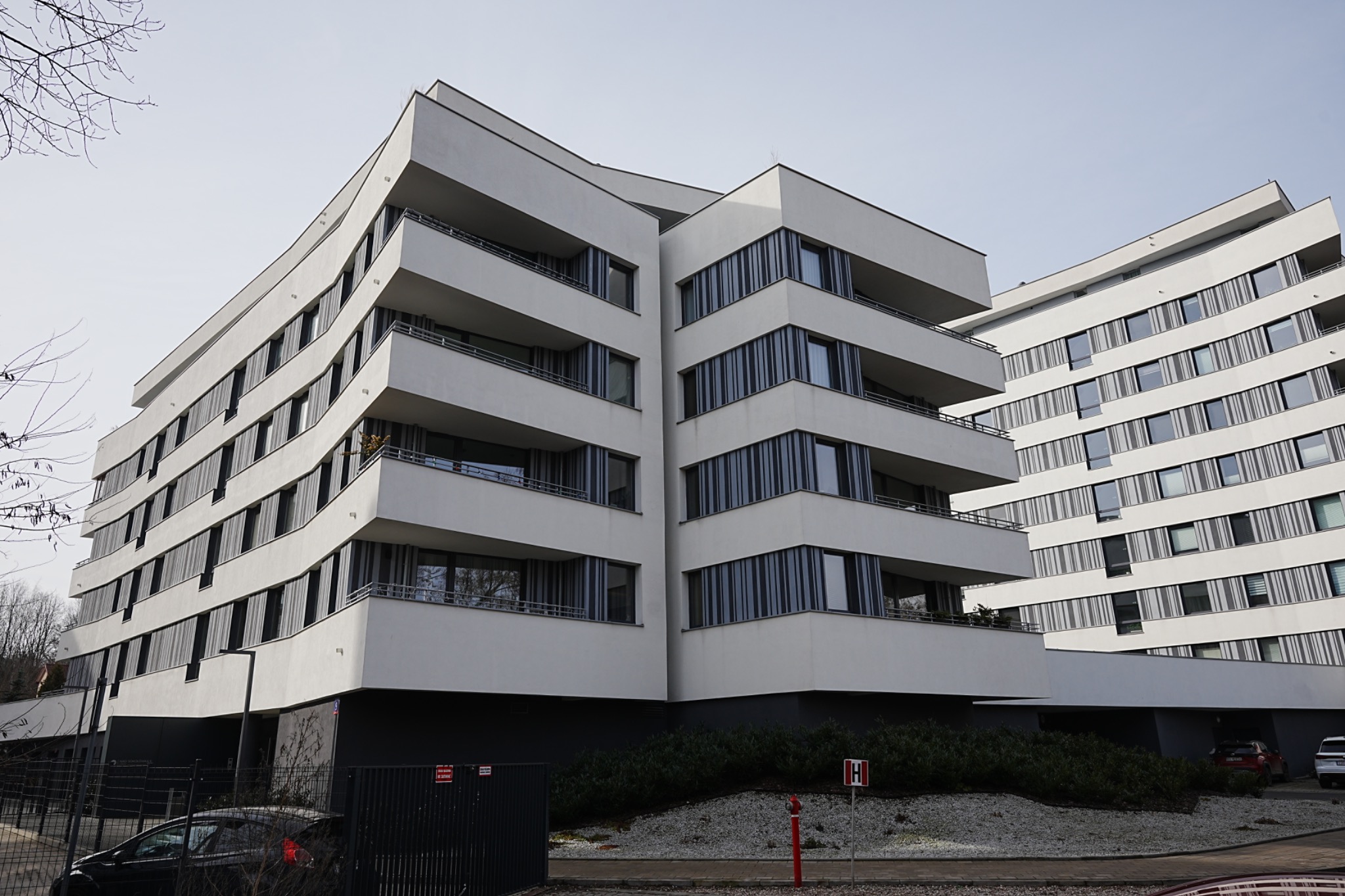 Nowoczesny, biały budynek wielorodzinny z balkonami i dużymi oknami, widoczny z dołu pod kątem. Minimalistyczna architektura, jasne tło nieba.