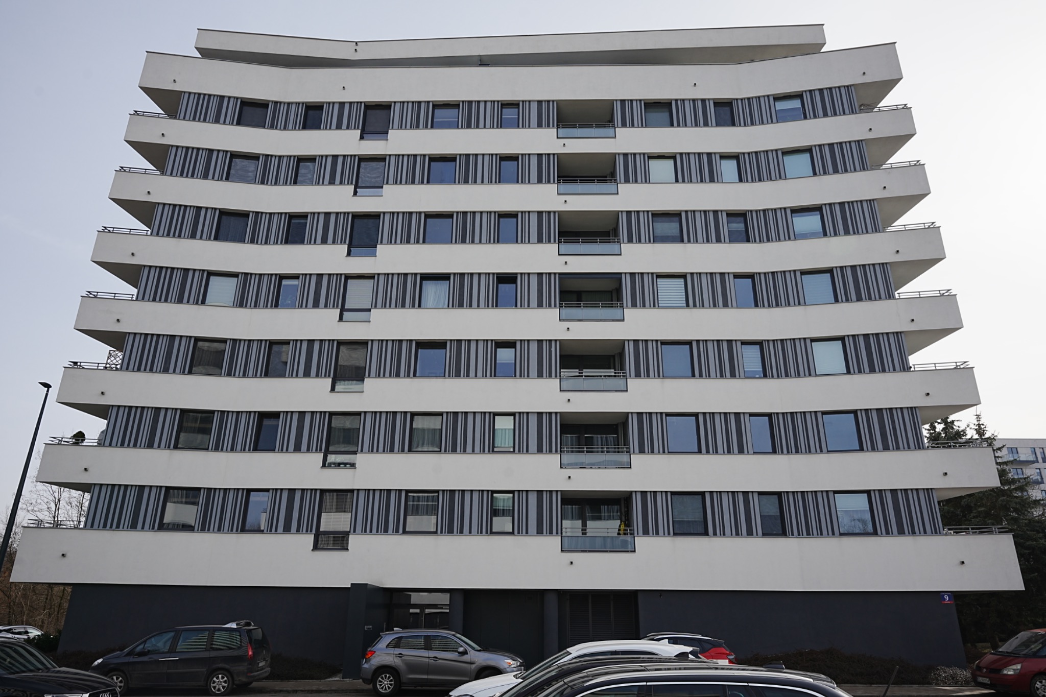 Nowoczesny budynek wielorodzinny z białymi, falującymi balkonami i szarymi pasami między oknami. Na dole widoczne zaparkowane samochody.