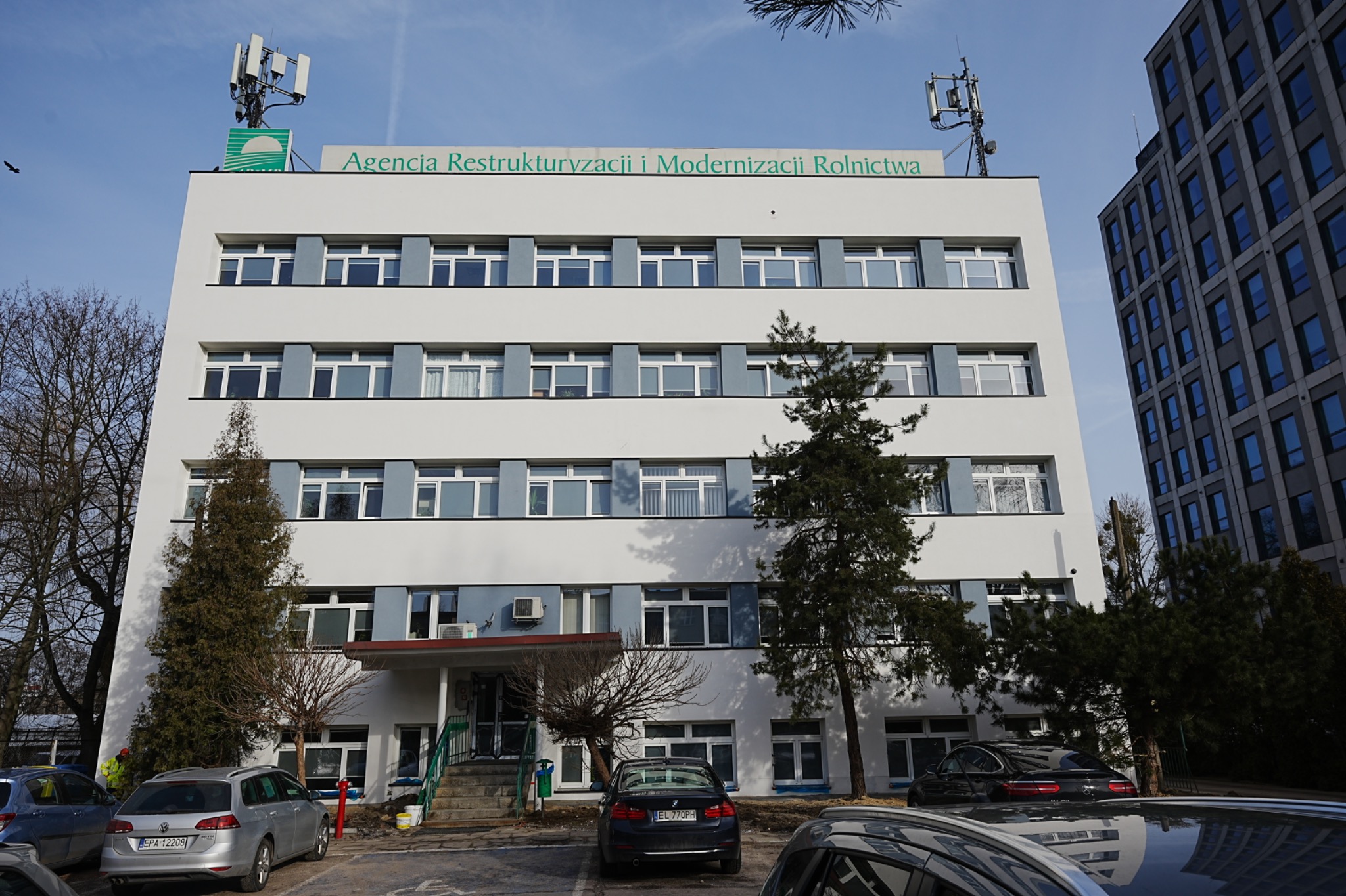 Biały budynek biurowy z szarymi akcentami okiennymi i logo Agencji Restrukturyzacji i Modernizacji Rolnictwa na dachu, otoczony drzewami i samochodami na parkingu.