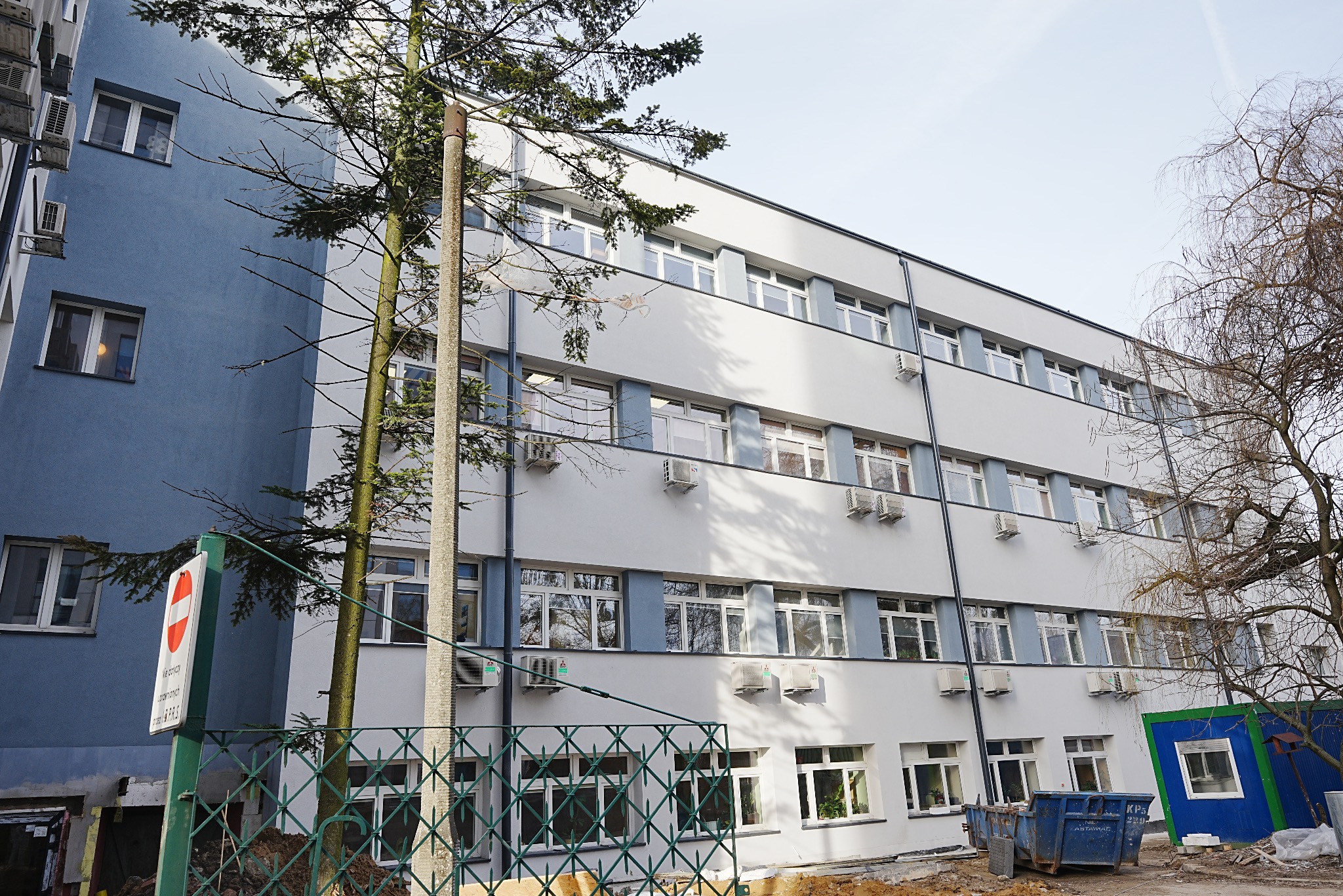 Odnowiony budynek biurowy z nową elewacją w odcieniach bieli i błękitu, widoczny z poziomu gruntu, z elementami zieleni i kontenerem na gruz w tle.