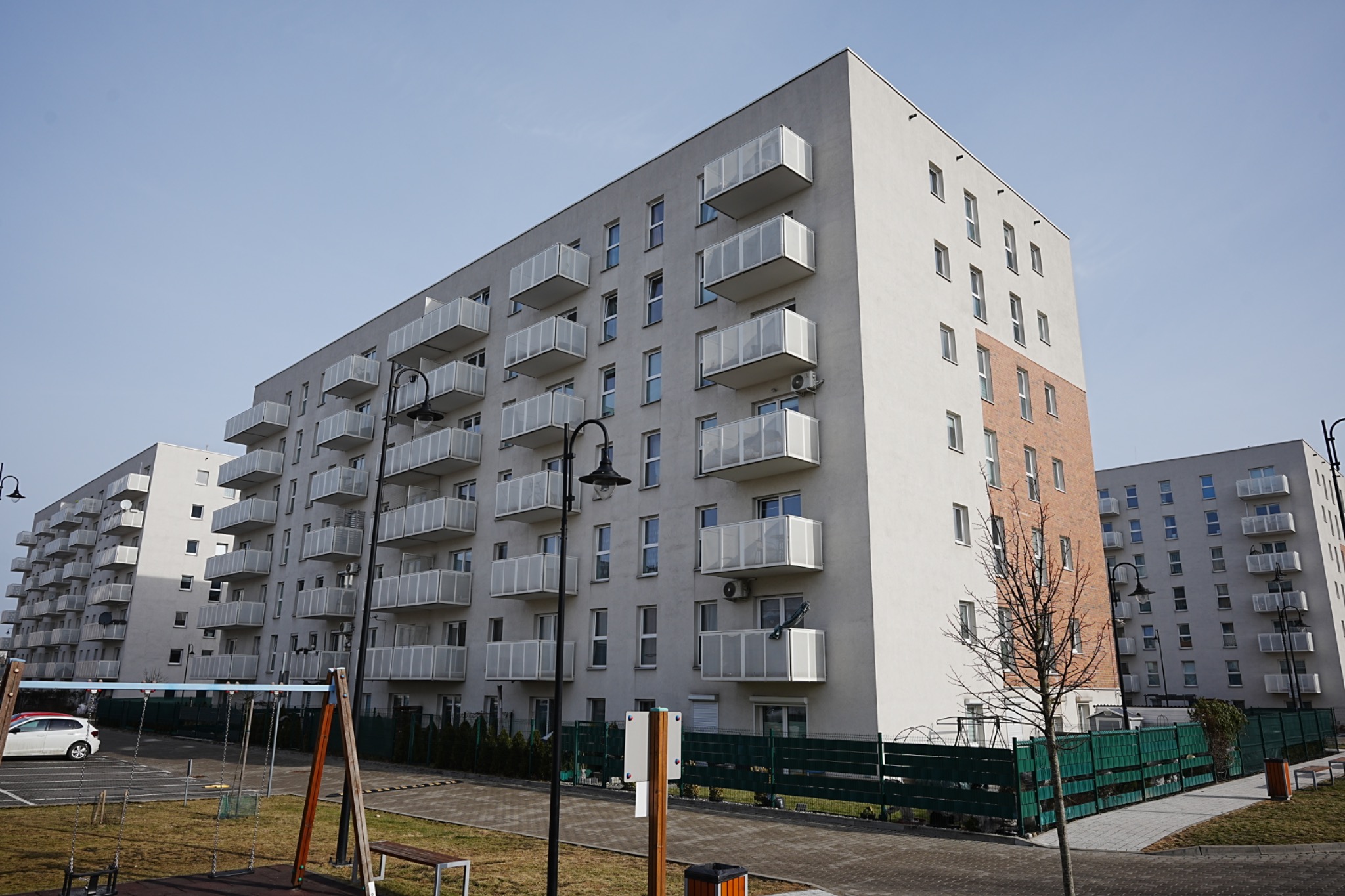 Nowoczesny, wielopiętrowy budynek mieszkalny z balkonami o białych balustradach. Część elewacji wykończona cegłą. Widok z poziomu chodnika, w tle inne budynki.