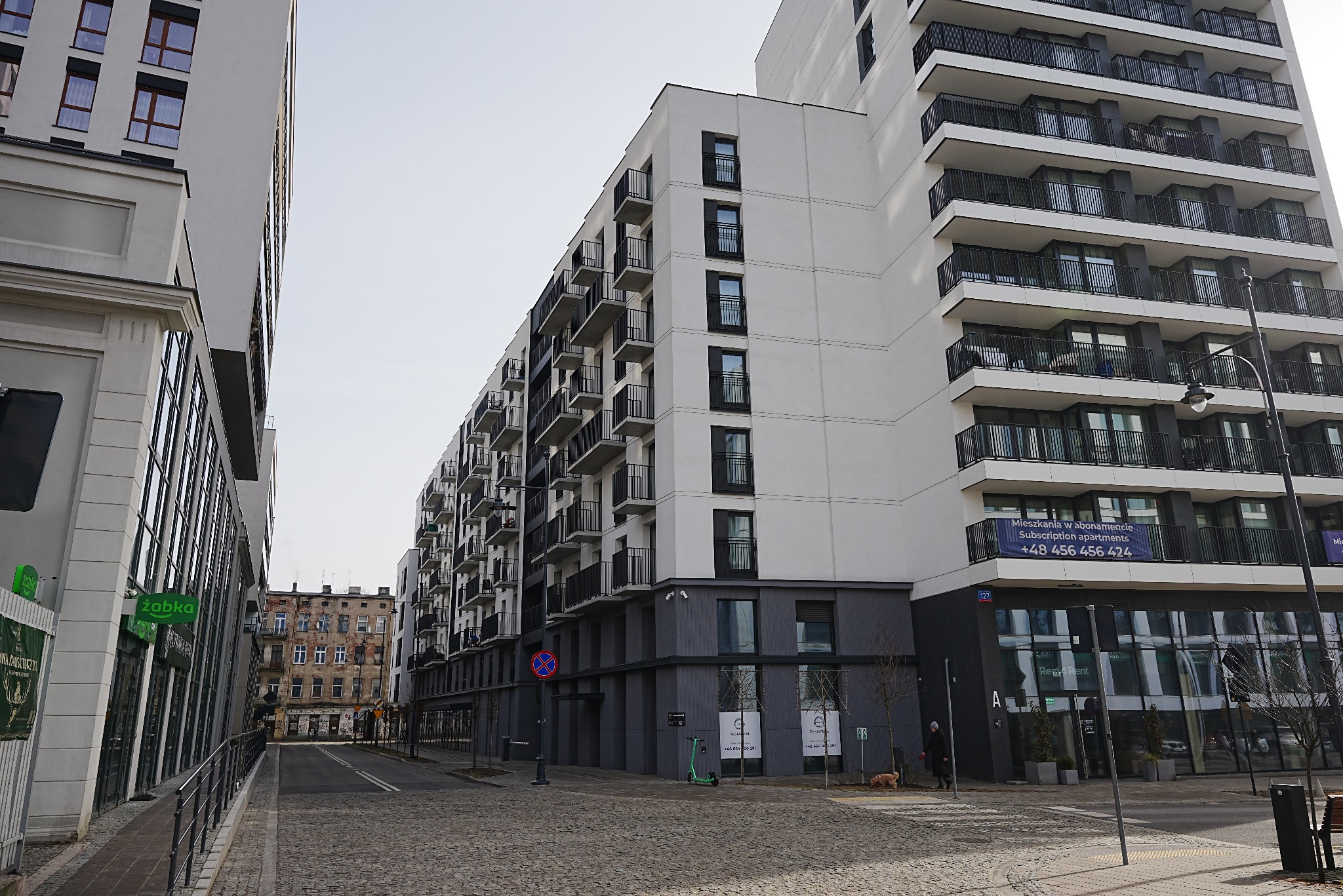 Nowoczesny budynek mieszkalny z balkonami, widoczny z perspektywy ulicy wyłożonej kostką. Kontrastujące kolory elewacji: biel i szarość. W tle starsza kamienica.