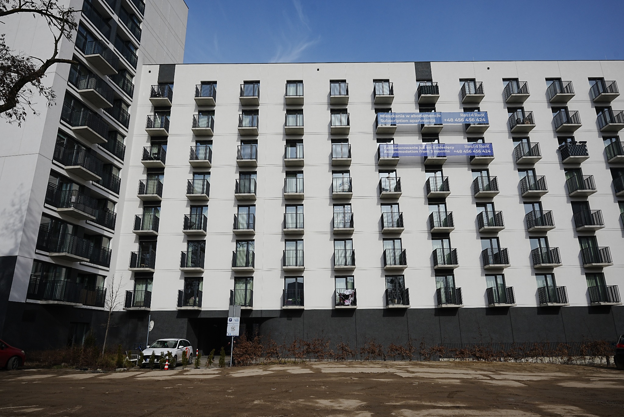 Nowoczesny budynek wielorodzinny w Łodzi z balkonami i szarą elewacją. Na niektórych balkonach wiszą banery reklamowe. Widok z dołu, w tle błękitne niebo.