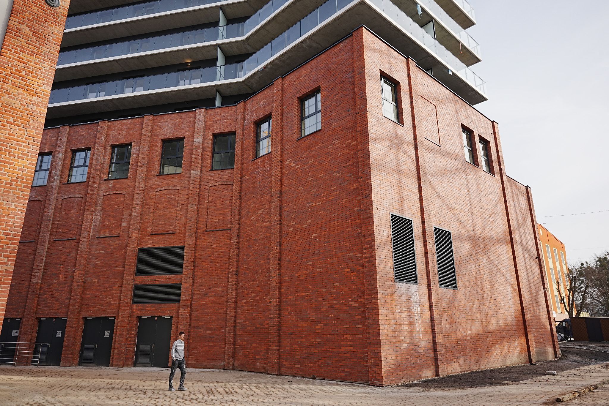 Ceglana fasada budynku z nowoczesną nadbudową. Widoczne okna, kratki wentylacyjne i chodnik z kostki. Osoba w oddali dodaje skali budowli. Architektura industrialna.