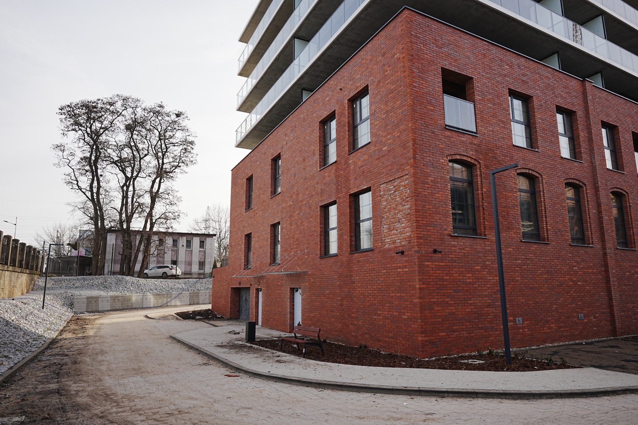 Ceglana elewacja budynku z oknami, widok z poziomu chodnika. W tle drzewa i fragment drugiego budynku. Nowoczesna architektura z elementami tradycyjnymi.